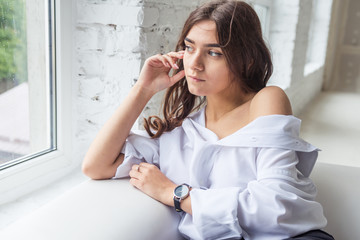 young cute sexy charming girl in a white man's shirt and dark jeans sitting by the window on a...