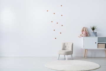 Pastel grey armchair next to wooden cabinet with books, toy and green plant in grey pot, copy space and golder stars stickers on empty white wall © Photographee.eu