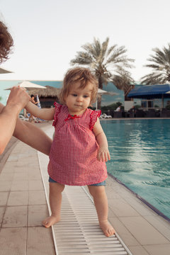 Funny little baby girl near swimming pool. Father holds infant by hands on pool background. Summer vacation with children outdoor.