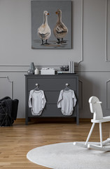 Little baby clothes hanging on grey wooden chest of drawers in elegant grey interior with wooden floor and molding on the wall