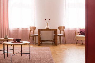 Chairs at wooden table with stylish vase with flowers in bright dining room interior with coffee tables on pink carpet