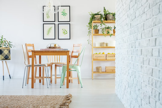 White brick wall in voguish dining room interior with wooden communal table and wooden and metal chairs, gallery of floral posters on the white empty wall