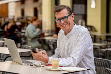 Fototapeta premium Happy attractive stylish mature man sending message on mobile while working on laptop outside cafe