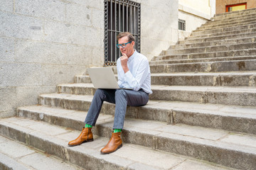 Fototapeta na wymiar Happy attractive mature man working on laptop checking email sitting on stairs outdoors urban area