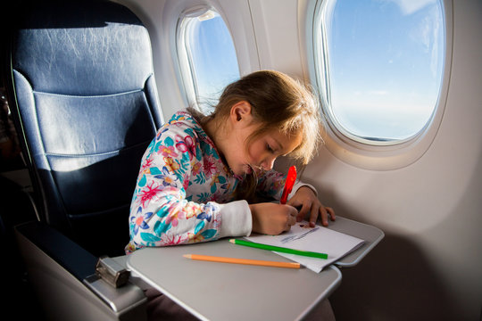 Child drawing picture with crayons in airplane. Little girl occupied while flying in aircraft. Travel with family and kids. Blue sky and sun outside the window