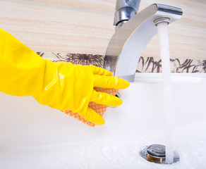 Female hand in yellow rubber glove washes white sink surface in the bathroom.