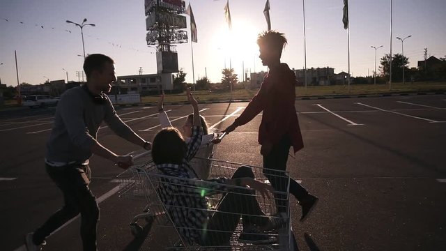 Young millennials people racing with shopping carts - Happy crazy friends having fun with trolleys in car park. Girls in the corts giving each other high five. Youth lifestyle, friendship and party