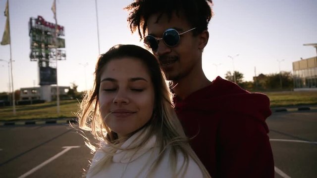Pretty couple hugging while standing outdoors on the parking zone. African man and caucasian woman standing emracing. Loving couple in evening lens flares