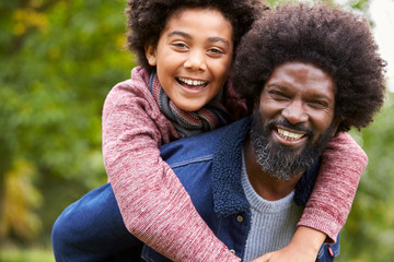 Black man piggybacking his pre-teen son in the park, both smiling to camera, close up