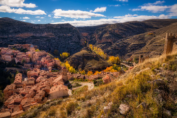 A landscape photo taken from above the town of Albarracin in Spain showing the ancient town, the surrounding mountains and the autumnal colors of the fauna under a bright blue sky with white clouds.