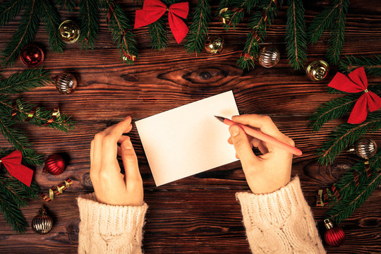 Female hands writing letter to Santa Claus on wooden background with christmas gifts and decoration top view. Vintage toned image with woman, wish list for christmas, flat lay, copy space, background.