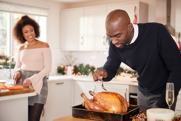 Young adult mixed race couple preparing Christmas dinner together at home, man basting roast turkey...