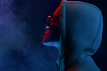 Neon profile portrait of young woman in sunglasses and hoodie. Studio shot with smoke in scene.