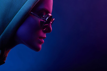 Neon profile portrait of young woman in sunglasses and hoodie. Studio shot with smoke in scene.
