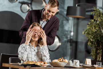 Handsome man closing eyes of his girlfriend to make surprise in restaurant