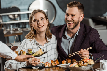 Smiling couple eating sushi rolls while waiter bringing new order in restaurant