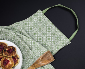 Green apron, wooden spatula and plate with cotton cheese pancakes on a dark background.