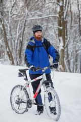 Determined young man standing next to a mountain bike, preparing for a ride in snow covered forest. Active sport lifestyle in cold weather.