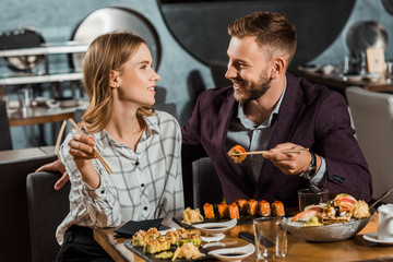 Happy young adult couple looking at each other while eating sushi rolls in restaurant
