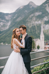 Naklejka premium A beautiful wedding couple walks on a background old cathedral, lake and mountains in a fairy Austrian town, Hallstatt.