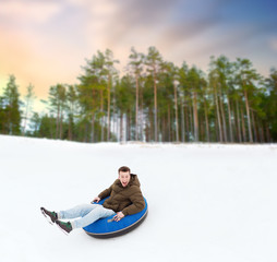 winter, leisure and entertainment concept - happy young man sliding down hill on snow tube over natural background