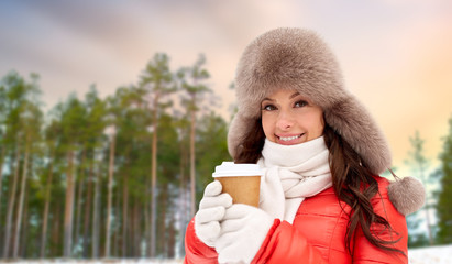 people, hot drinks and season concept - close up of happy woman in fur hat with coffee cup hat outdoors over winter forest background and snow