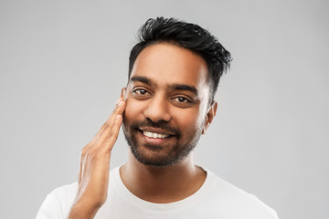 grooming, skin care and people concept - smiling young indian man touching his face over grey background