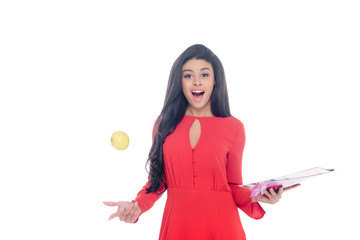 shocked african american girl in red dress holding magazine and throwing apple isolated on white