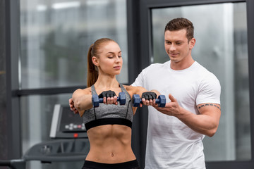 Naklejka premium handsome personal trainer helping sportswoman exercising with dumbbells in gym