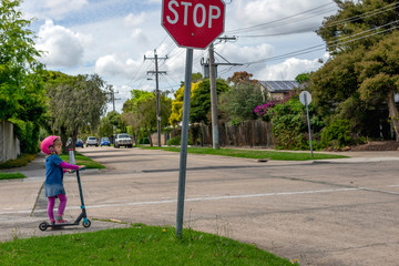 Young girl in pink outfit and pink helmet riding a kick-scooter, waiting at crossing with stop sign