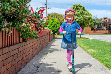 Young girl in pink outfit and pink helmet riding a kick-scooter on sidewalk