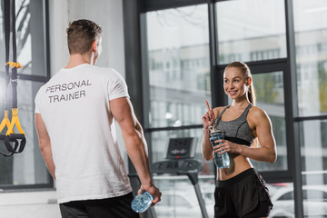 trainer and sportswoman holding sport bottles with water and looking at each other in gym