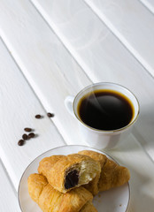 White cup of hot coffee with couple of croissants and chocolate.
