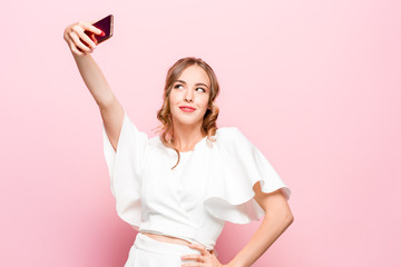 Portrait of a young attractive woman making selfie photo with smartphone on a pink studio background