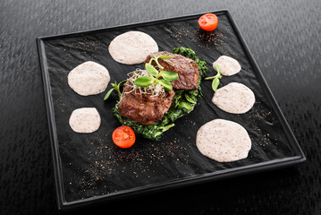 Medallions of veal on a spinach pillow with sprouts of grain, cream sauce and tomatoes on a black plate on a dark background. Beautiful serving of food. Flat lay. Space