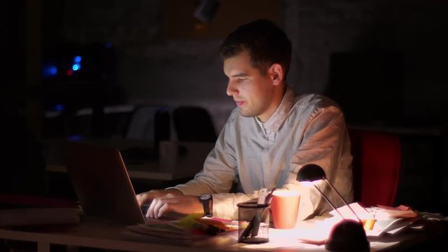 Handsome caucasian modern man is typing on laptop keyboard and turning off computer, finishing his work, late night and darkness in office