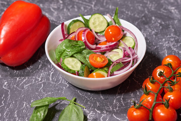 Vegetable salad in a white plate on a gray background, next to the Bulgarian pepper, tomatoes, spinach. Selective focus