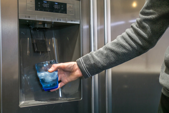 Male hand is pouring cold water and ice cubes from dispenser of home fridge.