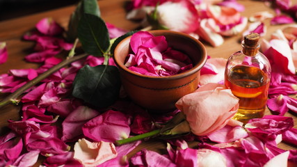 Clay bowl and aroma oil glass bottle among roses petals on the wooden table, natural raw material, selected focus