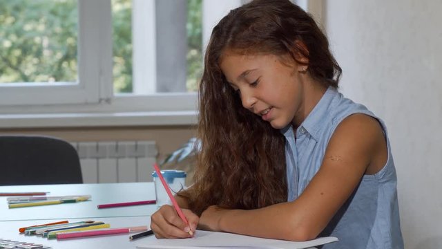 Adorable long haired girl enjoying drawing at school, smiling to the camera. Cute happy little schoolgirl coloring drawings in her sketchbook. Developing creativity in children concept.