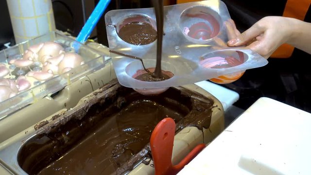 4K view of professional confectioners womans pours warm melted chocolate in plastic molds. 