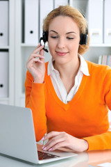 Young beautiful business woman in headset or call operator communicates by laptop computer. Orange sweater is uniform of a girl