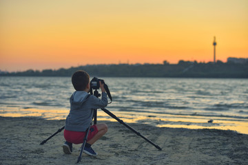 Creative child, kid photographer (a little boy) with a camera taking landscape pictures at sunset