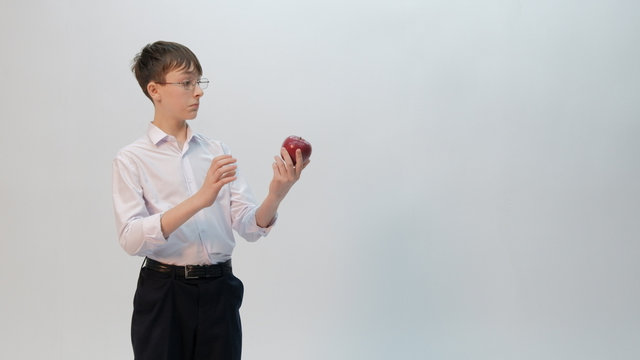 The teenager in glasses in astonishment looks at a red apple. Schoolboy guy in black pants and a white shirt with rolled up sleeves on a light background. Funny emotion in the image of a student.