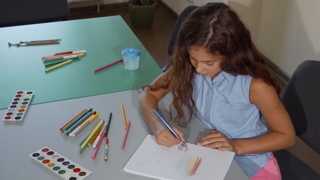 Young schoolgirl smiling to the camera while drawing. Top view shot of a long haired cute girl smiling cheerfully, whily coloring her drawings. Education, childhood, learning, leisure concept.