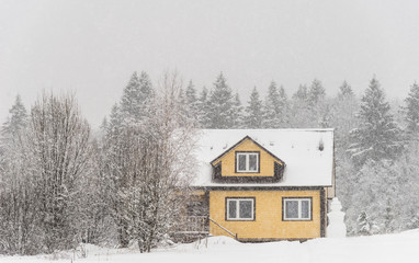 Yellow wooden house with terrace and snowman on forest background in heavy snow
