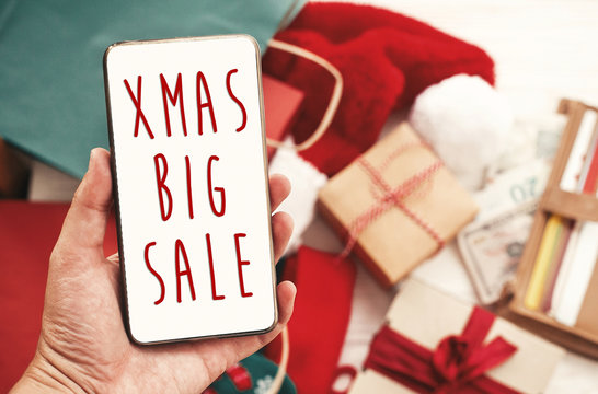 Christmas big sale text on phone screen, xmas sale sign. Special discount christmas offer sign. Hand holding phone with advertising message at credit cards, bags, clothes, gifts