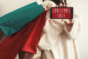 Christmas Sale text on phone screen. Special discount christmas offer sign. Big Sales. Girl holding phone with Advertising message and carrying shopping bags