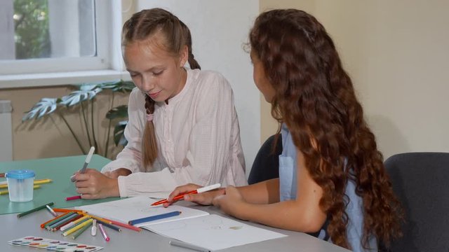 Two school friends talking and drawing together at art lesson. Cute red haired little girl and her friend enjoying coloring sketches. Schoolgirls working on art project. Children, friendship concept.