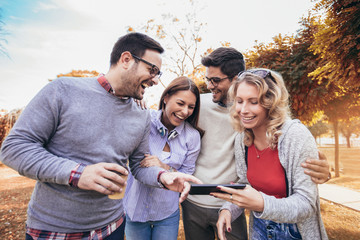 Four happy smiling young friends walking outdoors in the park holding digital tablet